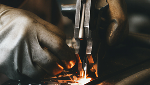 Close-up of a metalworker using a tool to cut metal, wearing protective gloves. Sparks fly amid scattered metal pieces on the worktable.