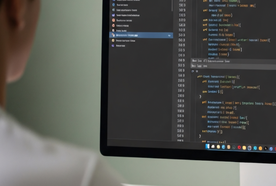 Person seen from behind viewing source code in a dark-themed editor on a large monitor above a white desk keyboard.