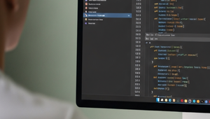 Person seen from behind viewing source code in a dark-themed editor on a large monitor above a white desk keyboard.