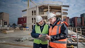 Two construction workers in safety gear review plans on a tablet at a building site with a partially constructed building and crane in the background.