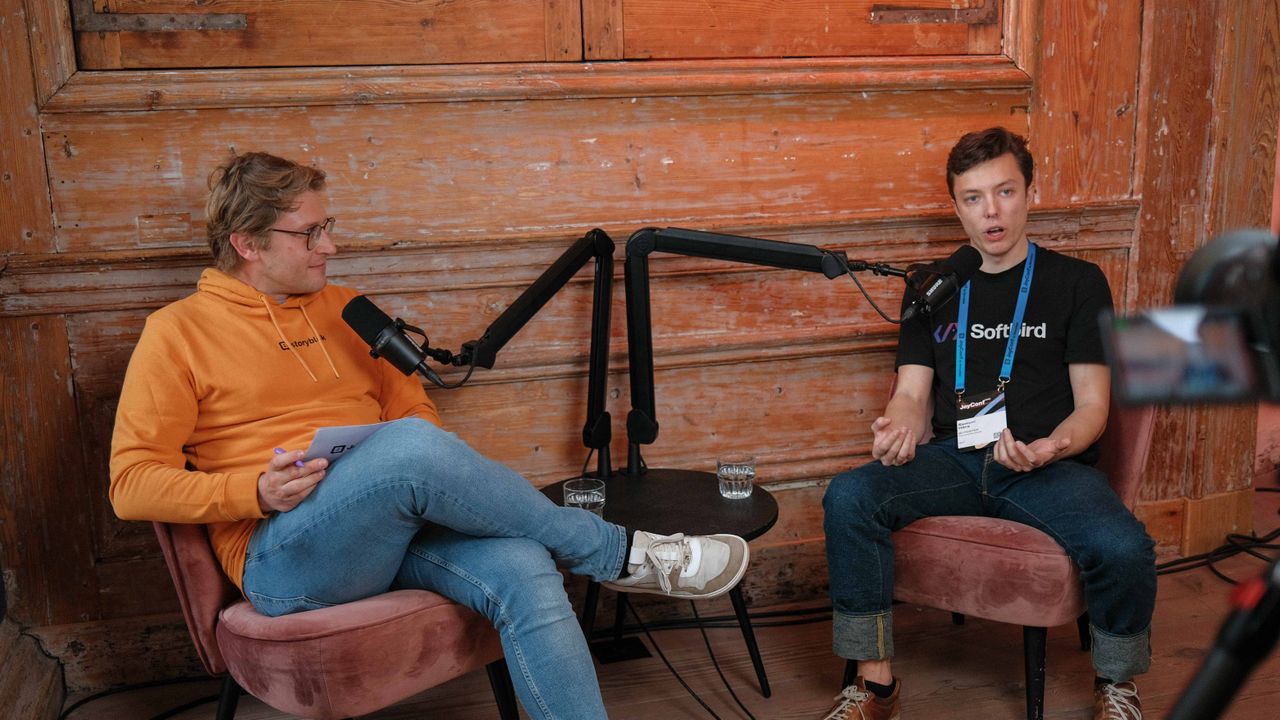 Two people are sitting in chairs, engaged in conversation with microphones, in a cozy room with a rustic wooden wall.