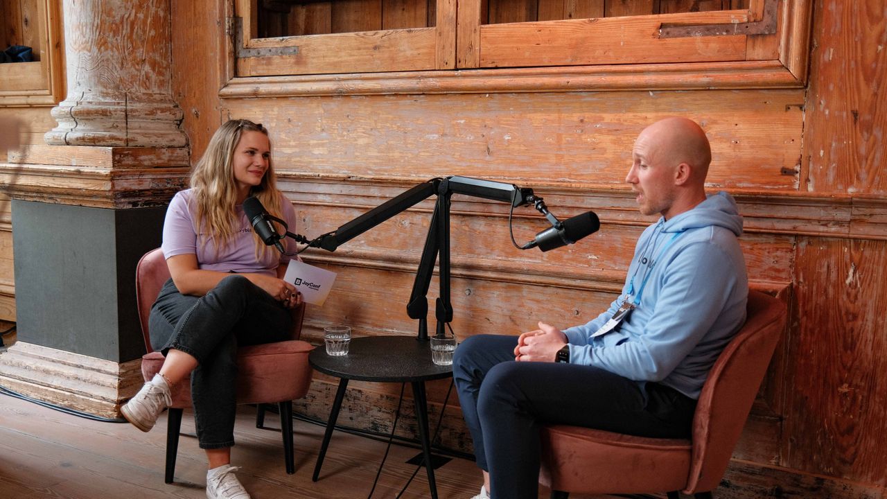 Two people sitting in chairs with microphones between them, engaged in conversation in a wood-paneled room.