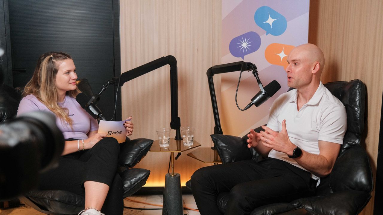 Two people sit in armchairs, engaged in a podcast discussion. They speak into microphones, with a colorful backdrop behind them.