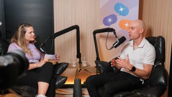 Two people sit in armchairs, engaged in a podcast discussion. They speak into microphones, with a colorful backdrop behind them.
