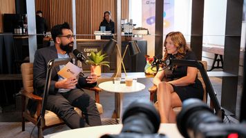 Two people with microphones sit in a cozy coffee bar setting, engaged in conversation.