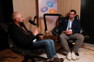 Two men sit in leather chairs recording a podcast-style conversation in a studio setup. The man on the left, bald with a beard, wears a black blazer, jeans, and brown boots, holding a notepad and speaking into a microphone. The man on the right wears glasses, a blue blazer, a white shirt, and gray pants, listening and preparing to respond. Multiple cameras and audio equipment are set up in the foreground, and a banner with colorful speech bubble icons hangs behind them.