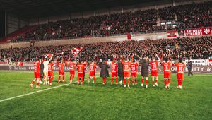 Football players in red kits applaud a packed stadium of supporters after a match, with fans waving red-and-white flags under the floodlights