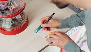 Person filling out a lotto ticket at a wooden counter, with a red container of scratch cards nearby.