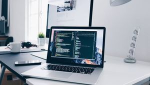 A sleek laptop with code on the screen is on a modern white desk, next to a monitor and other office items, in a bright workspace.