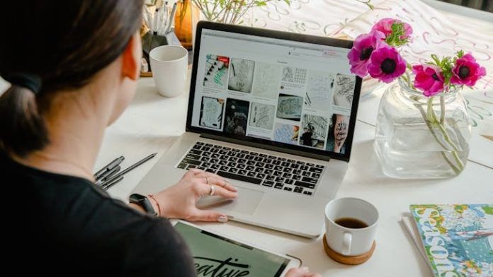 Person using a laptop displaying design images, with a tablet nearby. Flowers, a coffee cup, and a magazine are on the table.