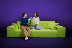 A man and woman sit facing each other on a lime-green sofa against a deep purple backdrop, both smiling as they hold a large green gift box wrapped with a purple "Pax" ribbon and a £50 gift tag between them. The man, wearing a navy sweater and tan trousers, sits with one leg crossed and white socks visible; the woman, in a white blouse and light-wash jeans, kneels on the cushions beside matching green pillows.