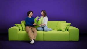A man and woman sit facing each other on a lime-green sofa against a deep purple backdrop, both smiling as they hold a large green gift box wrapped with a purple "Pax" ribbon and a £50 gift tag between them. The man, wearing a navy sweater and tan trousers, sits with one leg crossed and white socks visible; the woman, in a white blouse and light-wash jeans, kneels on the cushions beside matching green pillows.