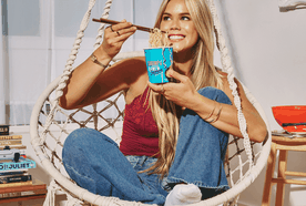 Woman sitting in a hanging chair, smiling while eating noodles from a cup with chopsticks, surrounded by books and a wooden floor.