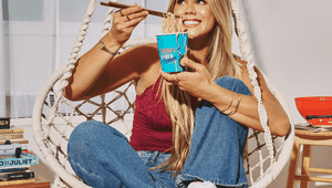 Woman sitting in a hanging chair, smiling while eating noodles from a cup with chopsticks, surrounded by books and a wooden floor.