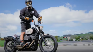 A motorcyclist wearing a helmet rides a black custom bike on an open road under a blue sky.