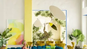 A bright, eclectic living room with high ceilings and exposed wooden beams painted yellow. A woman in a burgundy dress sits smiling on a large, plush teal modular sofa accented with green pillows. The space features colorful abstract art, large indoor plants, sculptural yellow chairs, and oversized circular hanging light fixtures. A sunlit courtyard with a tree is visible through a large open doorway, creating an airy, tropical atmosphere.