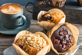 A selection of large, bakery-style muffins in brown paper wrappers arranged on a wooden surface. The muffins include varieties with chocolate chunks, crumble topping, and caramel pieces. Behind them sits a teal cup of cappuccino with latte art. The scene has a cozy, rustic café feel.