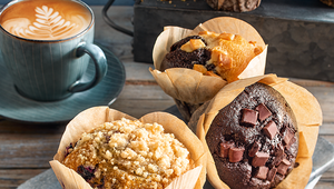 A selection of large, bakery-style muffins in brown paper wrappers arranged on a wooden surface. The muffins include varieties with chocolate chunks, crumble topping, and caramel pieces. Behind them sits a teal cup of cappuccino with latte art. The scene has a cozy, rustic café feel.