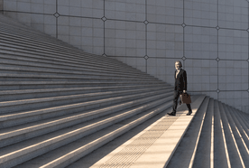 A man in a suit walking down a wide set of concrete steps, holding a brown briefcase, with geometric wall patterns in the background.