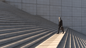 A man in a suit walking down a wide set of concrete steps, holding a brown briefcase, with geometric wall patterns in the background.