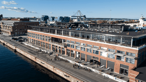 Aerial view of a waterfront industrial-style brick building with multiple balconies, beside a calm body of water under a clear blue sky.