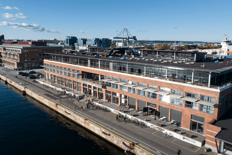 Aerial view of a waterfront industrial-style brick building with multiple balconies, beside a calm body of water under a clear blue sky.