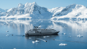 Cruise ship navigating icy waters with a backdrop of snowy mountains under a clear blue sky, small boats nearby.