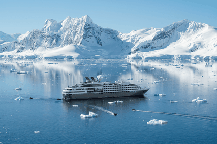 Cruise ship navigating icy waters with a backdrop of snowy mountains under a clear blue sky, small boats nearby.