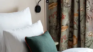 Cozy bed with white linens, green accent pillow, floral curtains, and a modern black wall lamp.