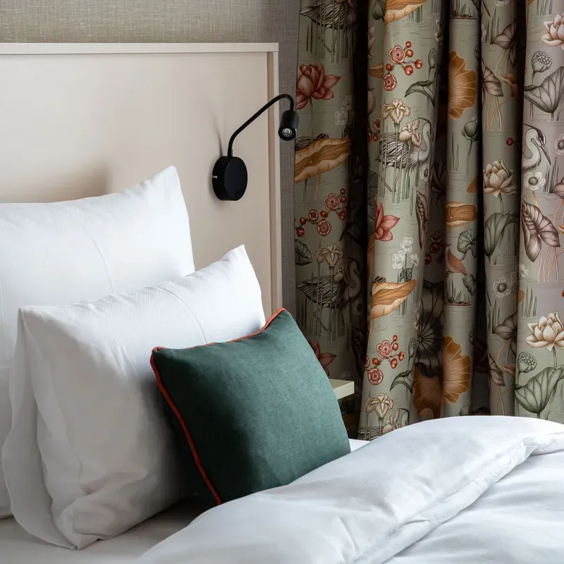 Cozy bed with white linens, green accent pillow, floral curtains, and a modern black wall lamp.