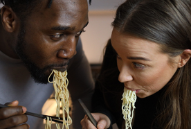 Two people eating noodles with chopsticks from white bowls, leaning towards each other, focused on their meal.