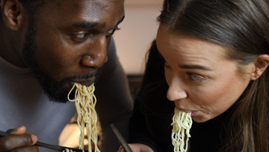Two people eating noodles with chopsticks from white bowls, leaning towards each other, focused on their meal.