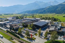 Aerial view of an industrial complex surrounded by greenery and mountains, with a parking lot filled with cars in the foreground.