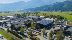 Aerial view of an industrial complex surrounded by greenery and mountains, with a parking lot filled with cars in the foreground.