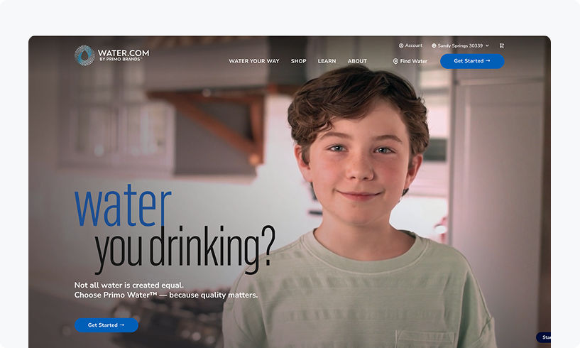 Young boy smiling in a kitchen, with text "water you drinking?" and "Choose Primo Water" on screen. Website navigation and account options visible.