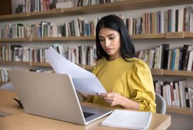 A woman in a yellow blouse sits at a desk in a library, reviewing papers beside an open laptop and notebook.