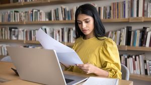 A woman in a yellow blouse sits at a desk in a library, reviewing papers beside an open laptop and notebook.