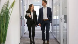 Two professionals in business attire walk down a modern office hallway, engaged in conversation, with glass doors on either side.