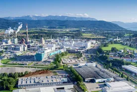 Aerial view of an industrial complex with smokestacks, surrounded by green fields and mountains under a clear blue sky.