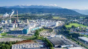 Aerial view of an industrial complex with smokestacks, surrounded by green fields and mountains under a clear blue sky.