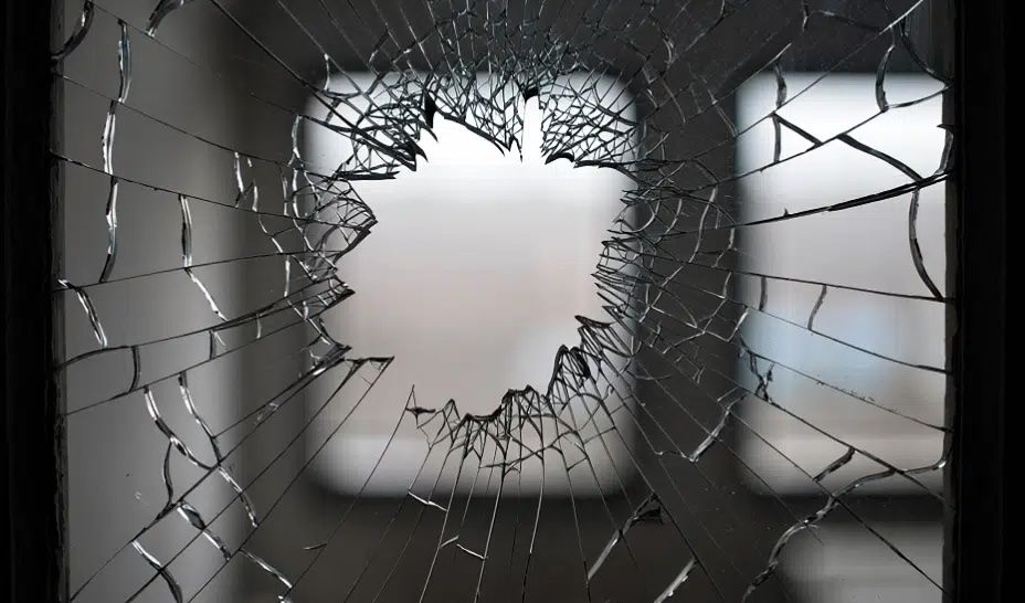 Close-up of a shattered window with a circular hole in the center, surrounded by radiating cracks. Blurred background visible through the glass.