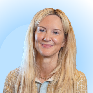 Smiling woman with long blonde hair wearing a plaid jacket over a light blue shirt against a soft blue background.
