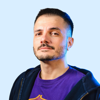 A man with short hair and a beard, wearing a purple t-shirt and a dark hoodie, looking at the camera against a light blue background.