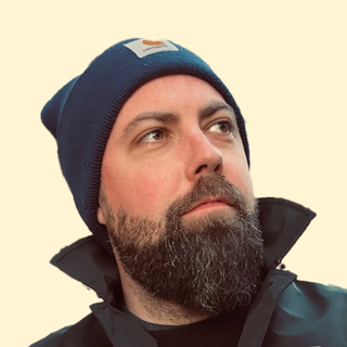 Bearded man wearing a blue knit beanie and black jacket, gazing upward against a neutral background.
