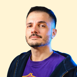 Man with a short haircut and beard, wearing a purple shirt and dark jacket, looking confidently at the camera against a beige background.
