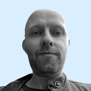 Black and white photo of a bald man with a short beard, wearing a buttoned jacket, against a light blue background.