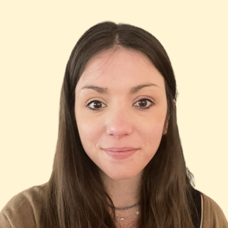 A woman with long brown hair and wearing a beige top, smiling slightly against a light background.