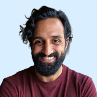 Smiling man with dark hair, beard, and maroon shirt against a light blue background.