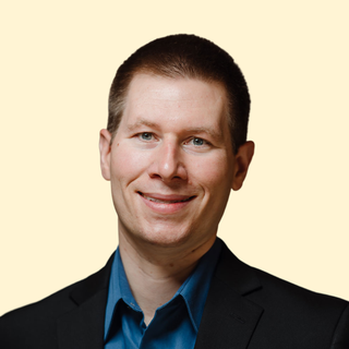 Smiling man in a black suit and blue shirt against a light beige background.