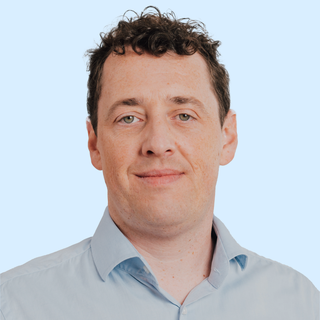 A man with curly brown hair wearing a light blue shirt, looking at the camera with a slight smile against a plain blue background.